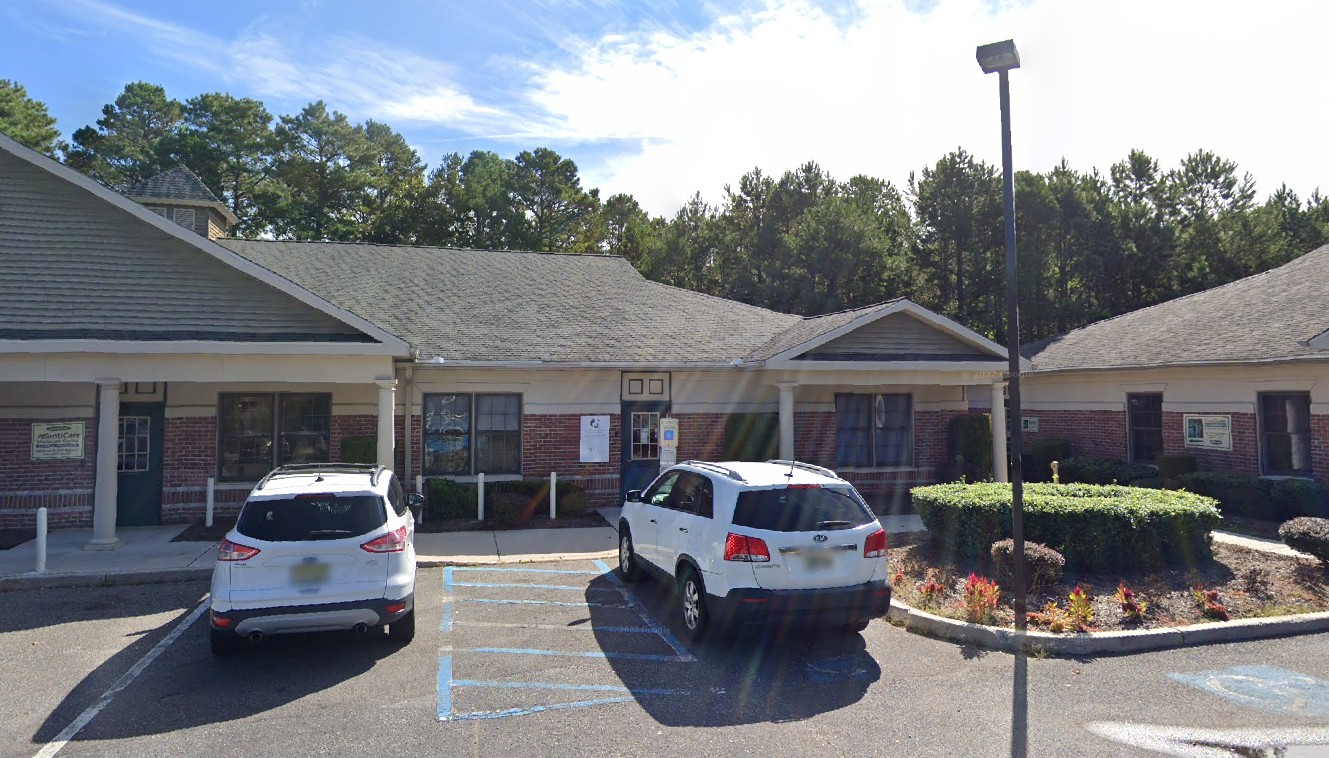 Exterior view of a medical office building with parked cars and landscaping in front. Pine trees in the background.